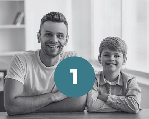 Foto di un uomo sorridente seduto e appoggiato a un tavolo sui 40 anni, magro e con una maglietta bianca. accanto a lui c'è un bambino sorridente con una camicia